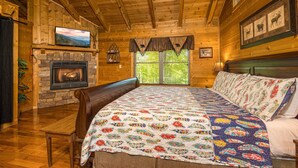A cozy log cabin room with a fireplace, a large bed, a television, and framed wildlife pictures on the wall.