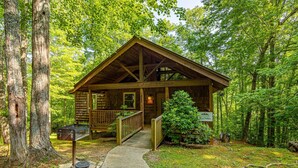 A rustic cabin with a wooden deck, a covered porch, and a small front yard with a mailbox and a sign.