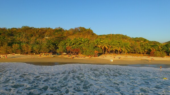 A beach with clear waters, palm trees, and a hilly backdrop.