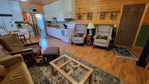 A cozy living room with a wooden floor, a coffee table, and two armchairs. A kitchen with white cabinets and a stove is visible in the background.