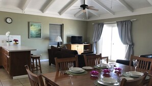 Un comedor con una mesa de madera dispuesta para cuatro personas, un sofá, un televisor y una zona de cocina con un reloj y flores en la encimera.
