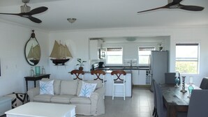 A spacious living area with a sofa, coffee table, and dining table. A kitchen with a refrigerator, sink, and window is visible in the background.