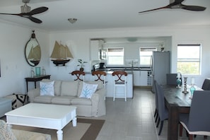 A spacious living area with a sofa, coffee table, and dining table. A kitchen with a refrigerator, sink, and window is visible in the background. - Trade Winds - Oceanview, Private Pool, Short Walk To Pink Sand Beach, Central. (Governors Harbour)