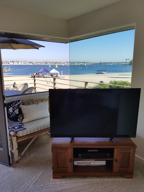 Een kamer met een grote televisie op een houten standaard, een bank met kussens, en uitzicht op een strand met boten.