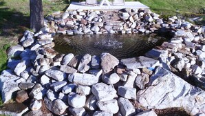 A circular stone structure with a small water feature in the center, surrounded by rocks and two white chairs with a table. - Quiet Peaceful Carmi Mountain Home. (Penticton)