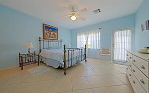Une chambre à coucher comprenant un lit, un ventilateur de plafond, une fenêtre avec des rideaux, une chaise et une commode.