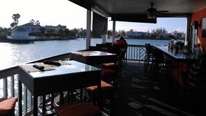 An outdoor bar area with tables and chairs, overlooking a body of water with buildings in the background.