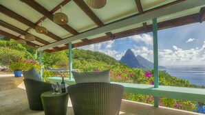 Un área de estar al aire libre con techo, mobiliario de mimbre, una botella de vino y vistas a un paisaje montañoso y al océano.