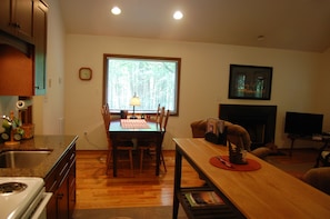 A dining area with a wooden table, chairs, a window with blinds, a clock, and a television in the background. - Deer Run - Mountain Getaway (Montebello)