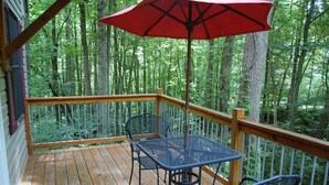 Um deck de madeira com mesa e cadeiras de metal, cercado por guarda-corpo metálico, com vista para uma área arborizada.
