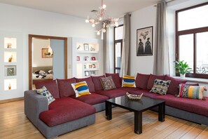 Un salon moderne avec un canapé d’angle gris, une table basse noire et un lustre. On y trouve des étagères avec des tableaux encadrés et une grande fenêtre avec des rideaux.