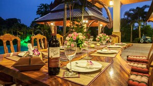 Un espace extérieur aménagé pour les repas, avec une table en bois dressée pour un repas, une bouteille de vin et un gazebo en arrière-plan.