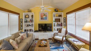 Una sala de estar con sofá, sillón, televisor, estantería y ventilador de techo.