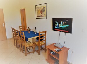 Ruokailutila, jossa on neljälle katettu ruokapöytä, seinälle asennettu televisio ja television alla oleva puinen kaappi.