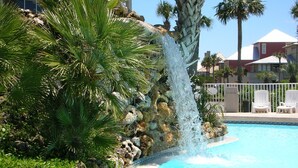 Une piscine agrémentée d’une cascade, entourée de palmiers et d’un mur de pierre.
