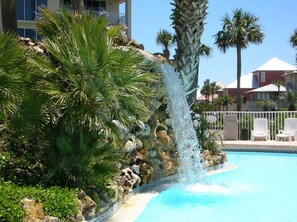 Une piscine agrémentée d’une cascade, entourée de palmiers et d’un mur de pierre.