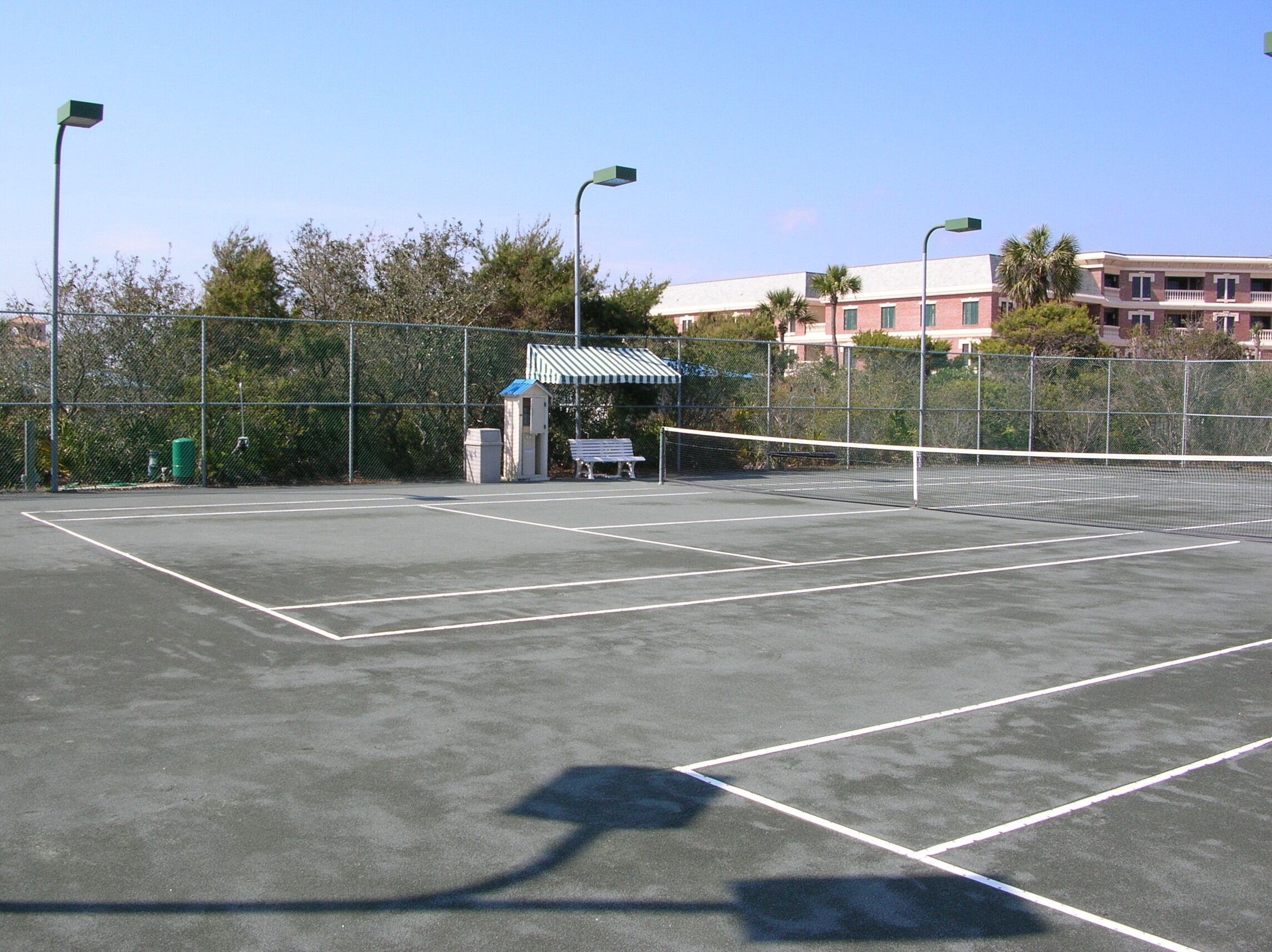 An outdoor tennis court with a fence, a small pavilion, and buildings in the background.