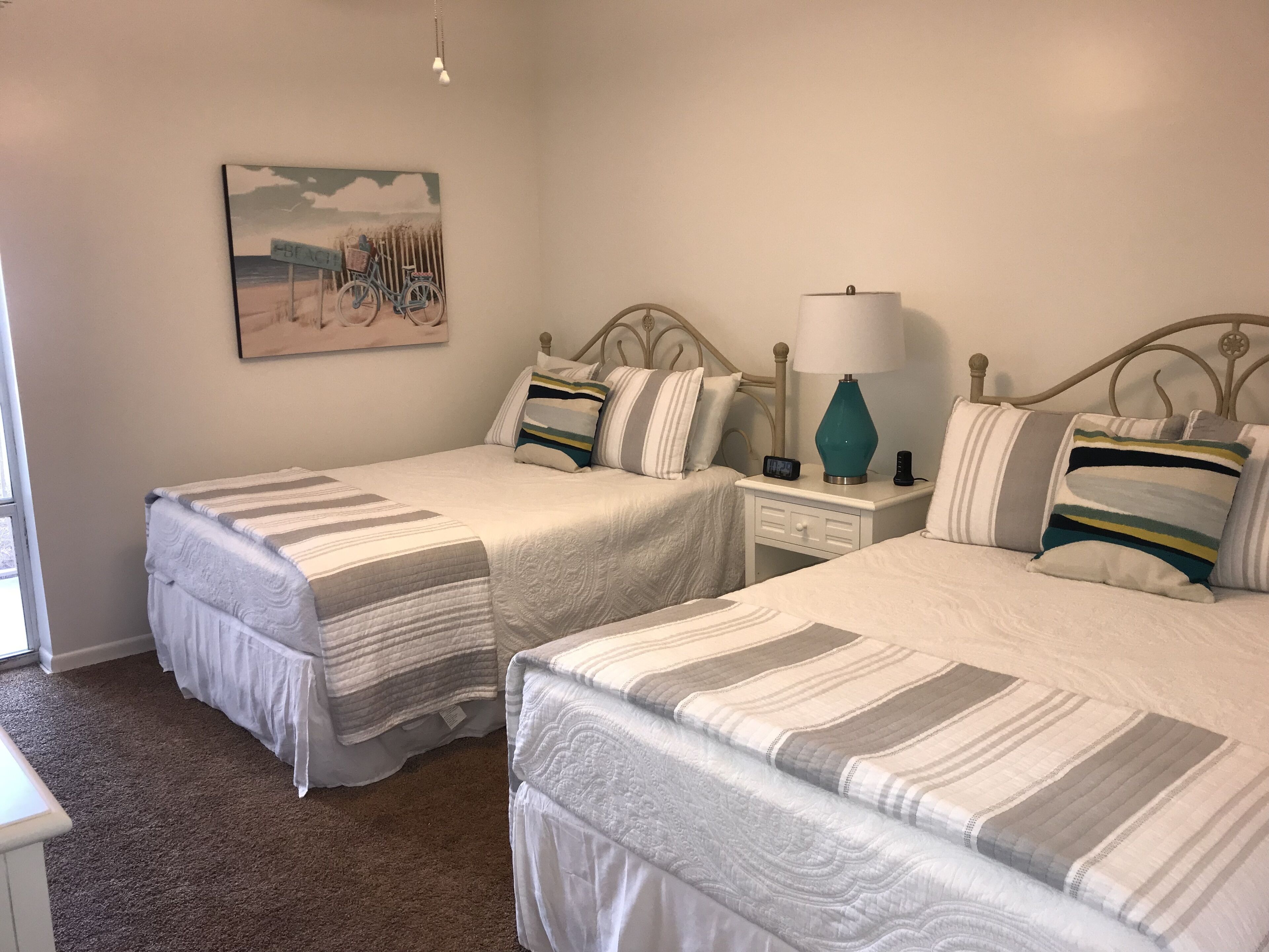 A hotel room with two single beds, a nightstand with a lamp, a painting on the wall, and a carpeted floor.