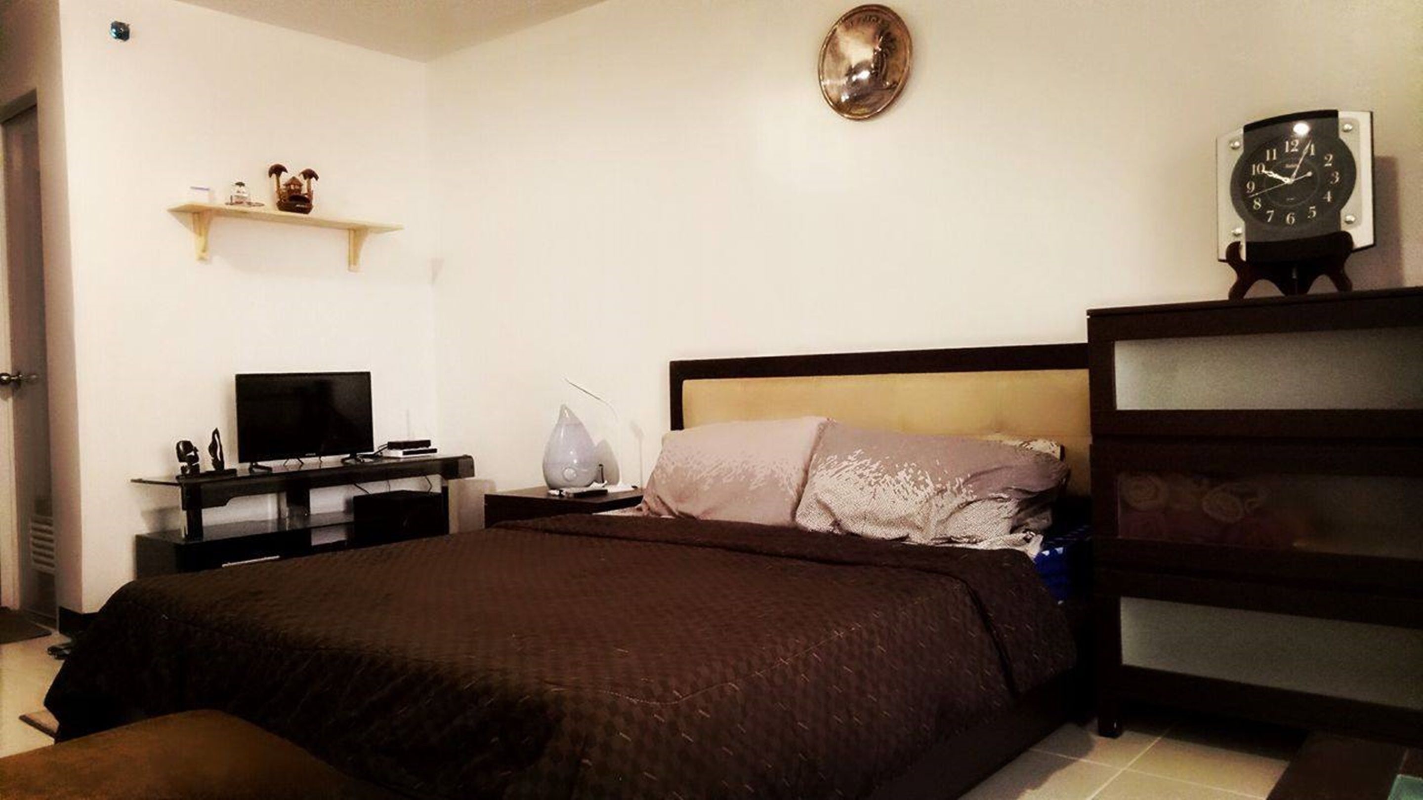 A bedroom with a bed, a TV on a stand, a bedside table with a lamp, and a clock on the wall.