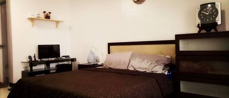 A bedroom with a bed, a TV on a stand, a bedside table with a lamp, and a clock on the wall.