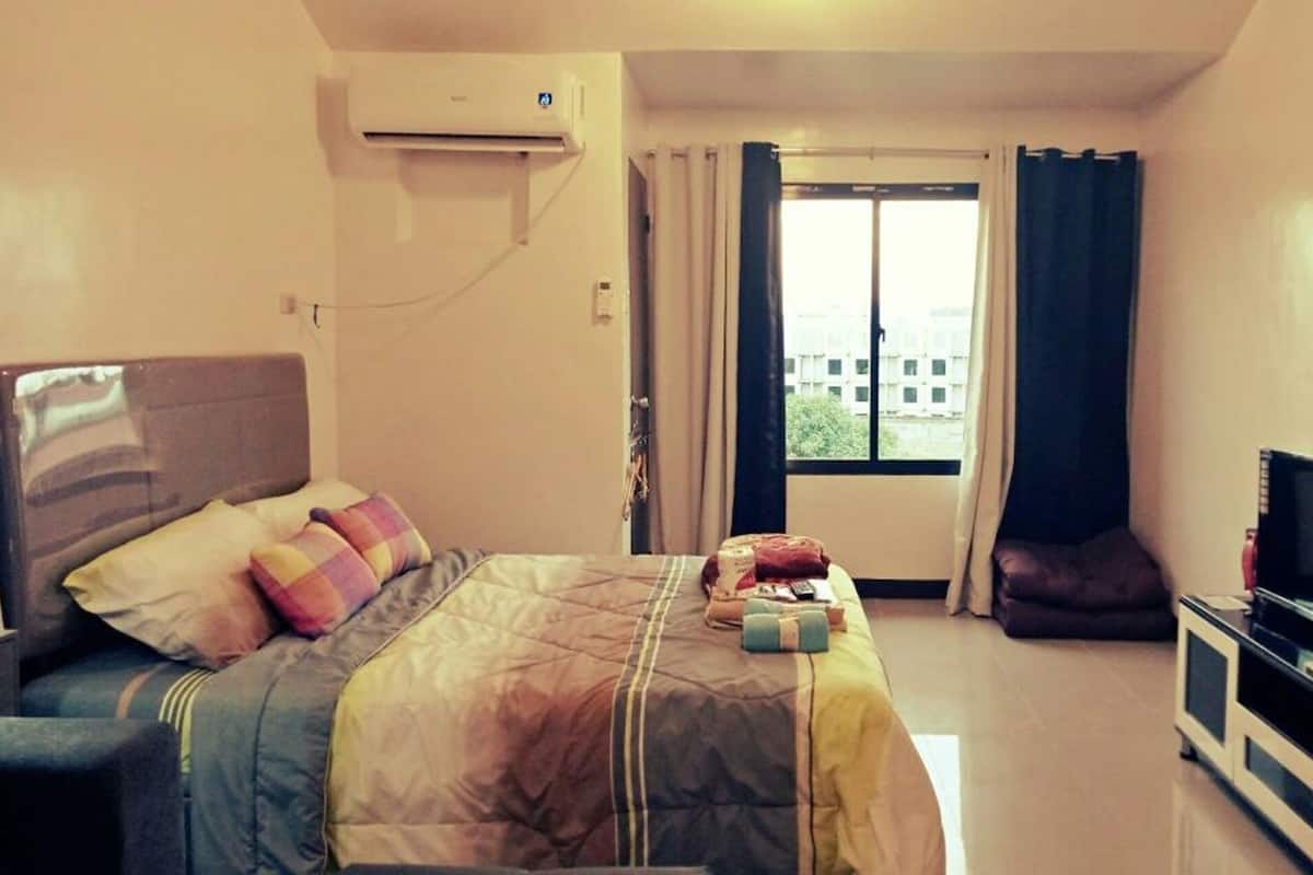 A hotel room with a bed, a TV on a stand, a window with curtains, and an air conditioning unit.