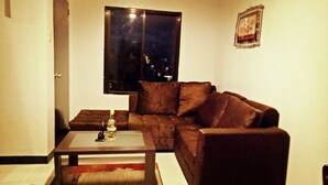 Ein Wohnzimmer mit einem braunen Sofa, einem Glastisch und einem kleinen Tisch mit einer Flasche und einem Dekorationsgegenstand.