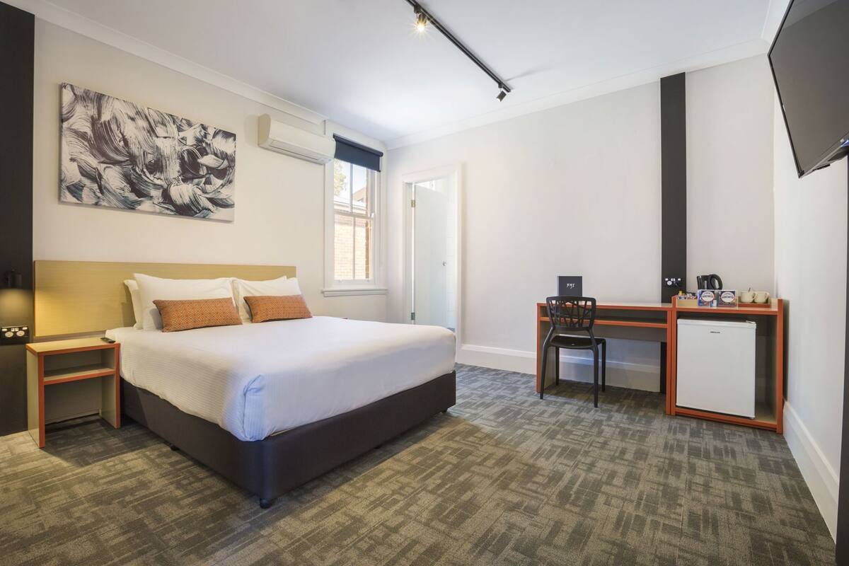 Habitación de hotel con cama, escritorio, silla, refrigerador pequeño y televisor montado en la pared.