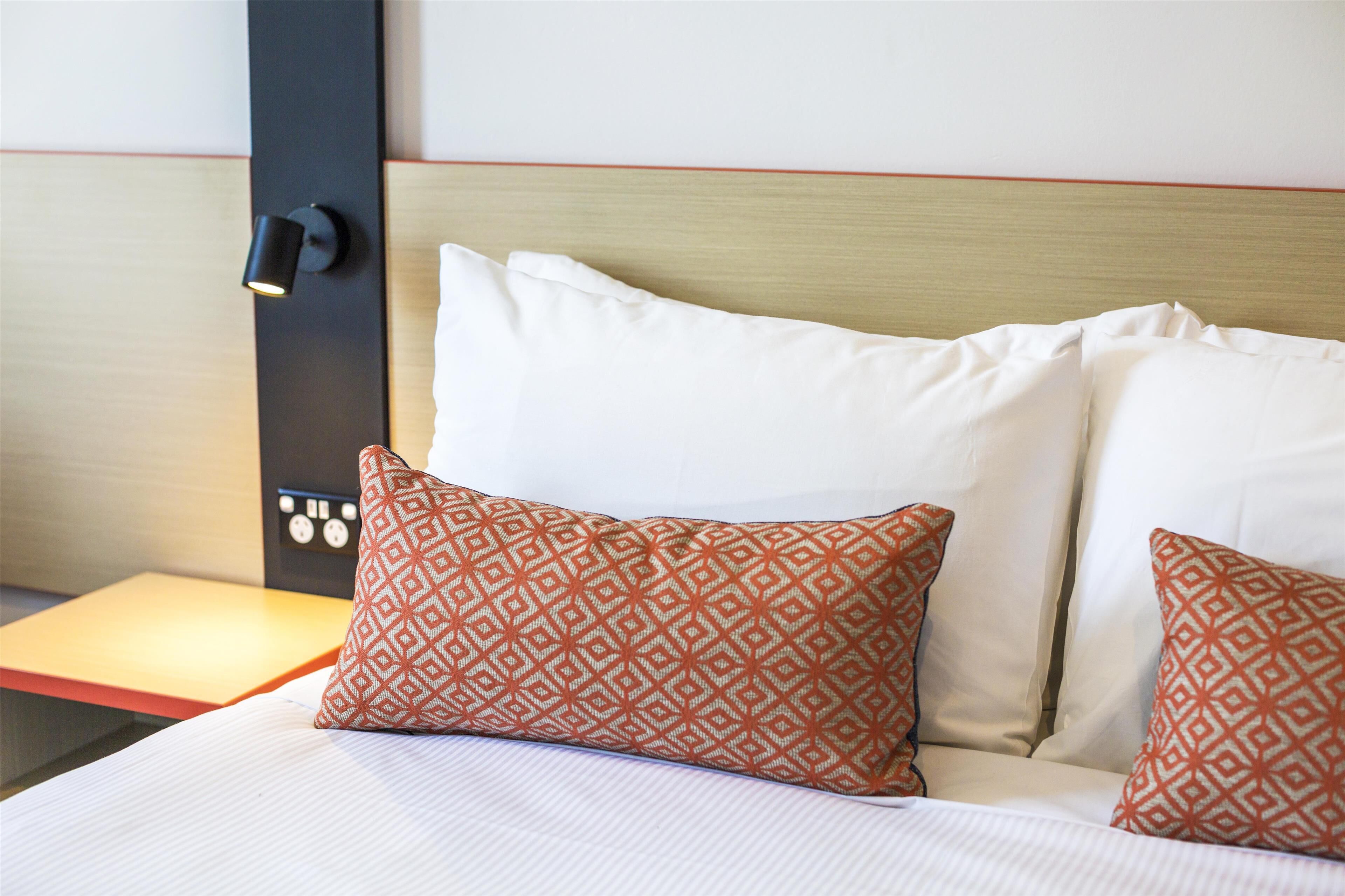 A neatly made bed with white linens and two patterned pillows, a bedside table with a lamp, and a wall-mounted control panel.