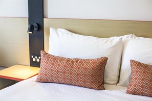 A neatly made bed with white linens and two patterned pillows, a bedside table with a lamp, and a wall-mounted control panel.