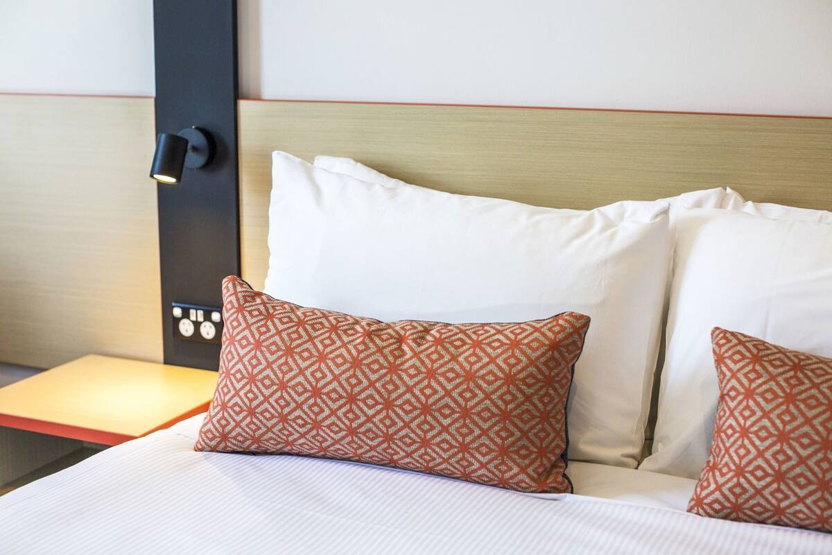 Habitación de hotel con cama, dos almohadas estampadas, una mesita de noche y un panel de control en la pared.