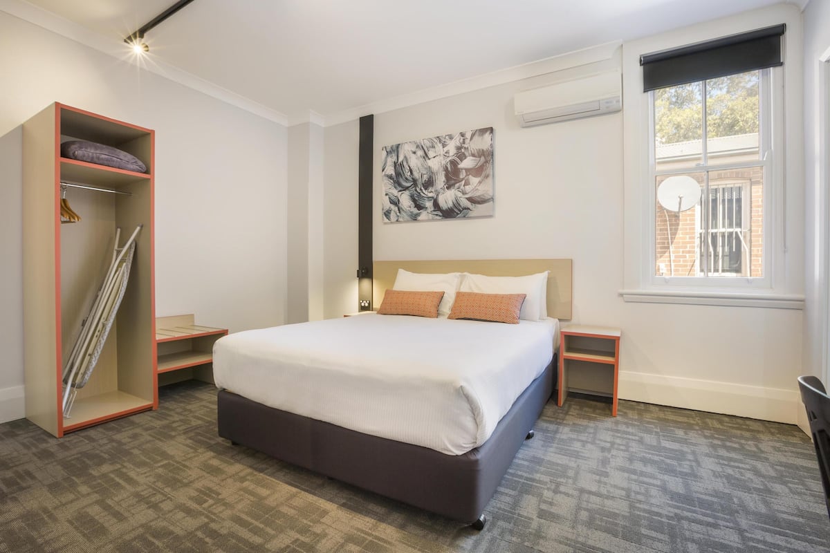 Una habitación de hotel moderna con una cama grande, un armario, una mesita y una ventana con vistas al exterior.
