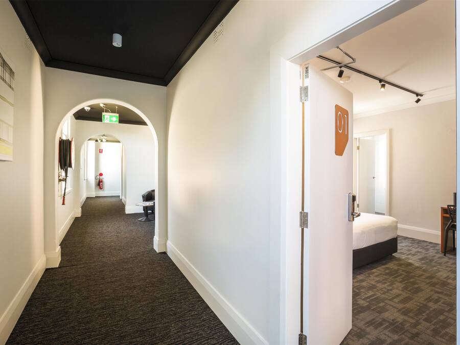 A hotel corridor with a carpeted floor, a bed in a room with a headboard, and a door with a number sign.