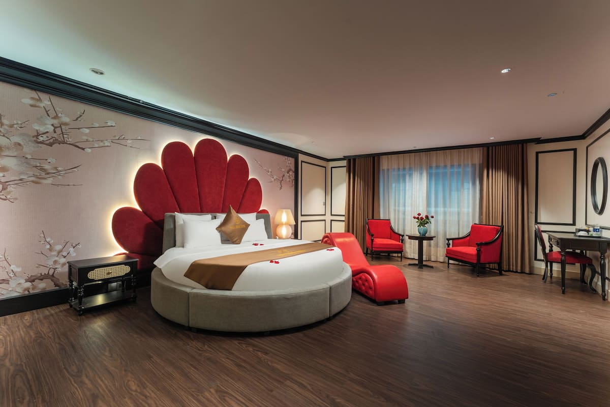 Una habitación de hotel moderna con una cama redonda, sillones tapizados en rojo, una mesita con un florero y un espejo montado en la pared.