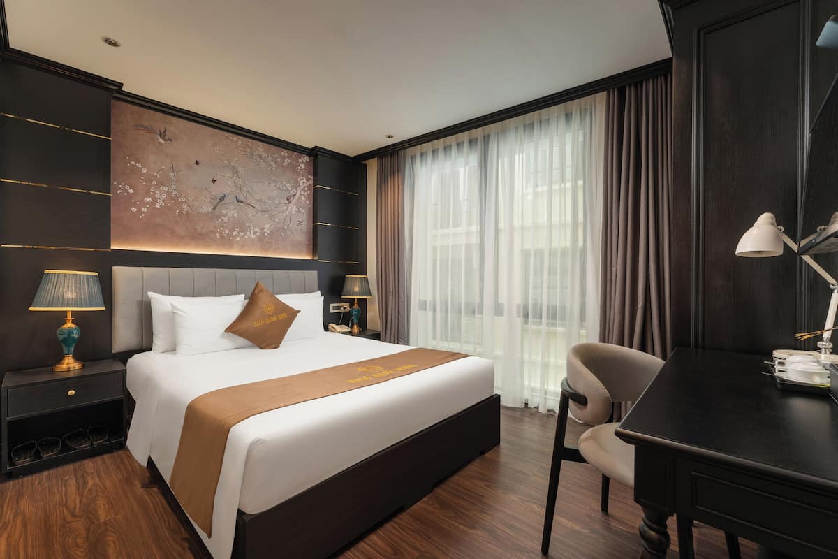 Habitación de hotel con una cama grande, un escritorio y una silla. Tiene una ventana con cortinas, una obra de arte en la pared y una mesita de noche con una lámpara.