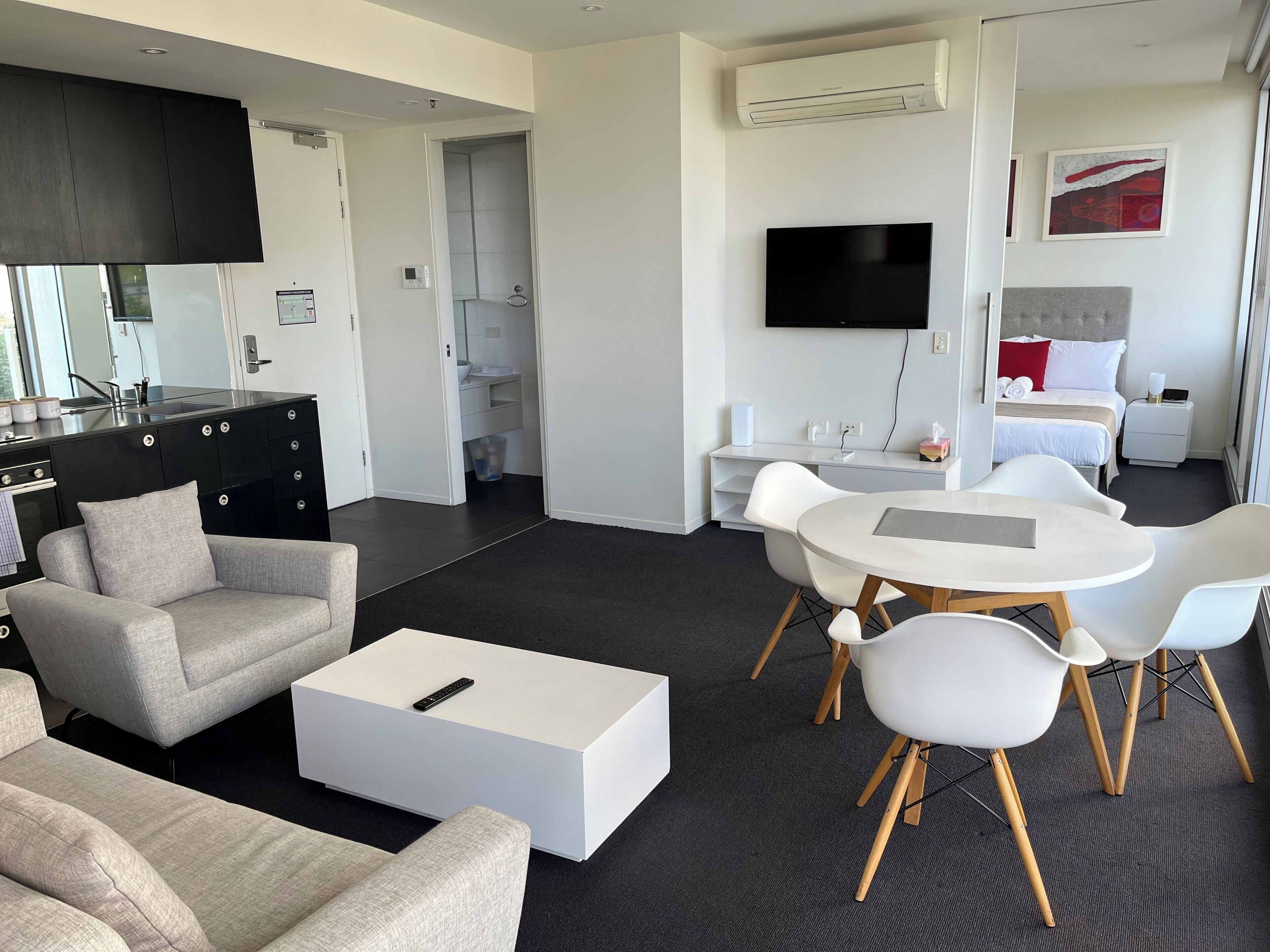 A modern hotel room with a kitchenette, a dining area with white chairs, a sofa, and a flat-screen TV mounted on the wall.