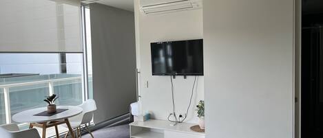 Quarto de hotel moderno com uma janela grande, uma TV de tela plana na parede, uma mesa de consola branca e uma mesa redonda com cadeiras.