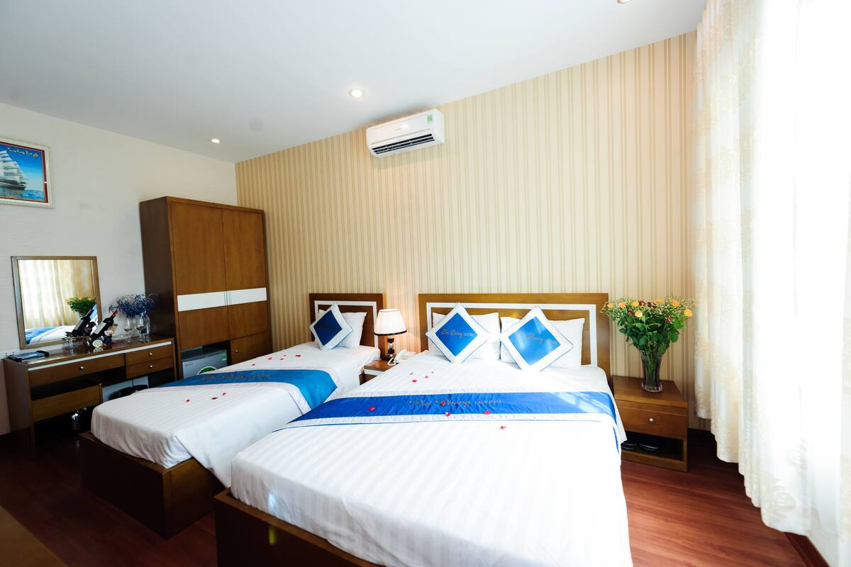 A hotel room with two beds, a wooden wardrobe, a desk with a lamp, and a wall-mounted air conditioner.