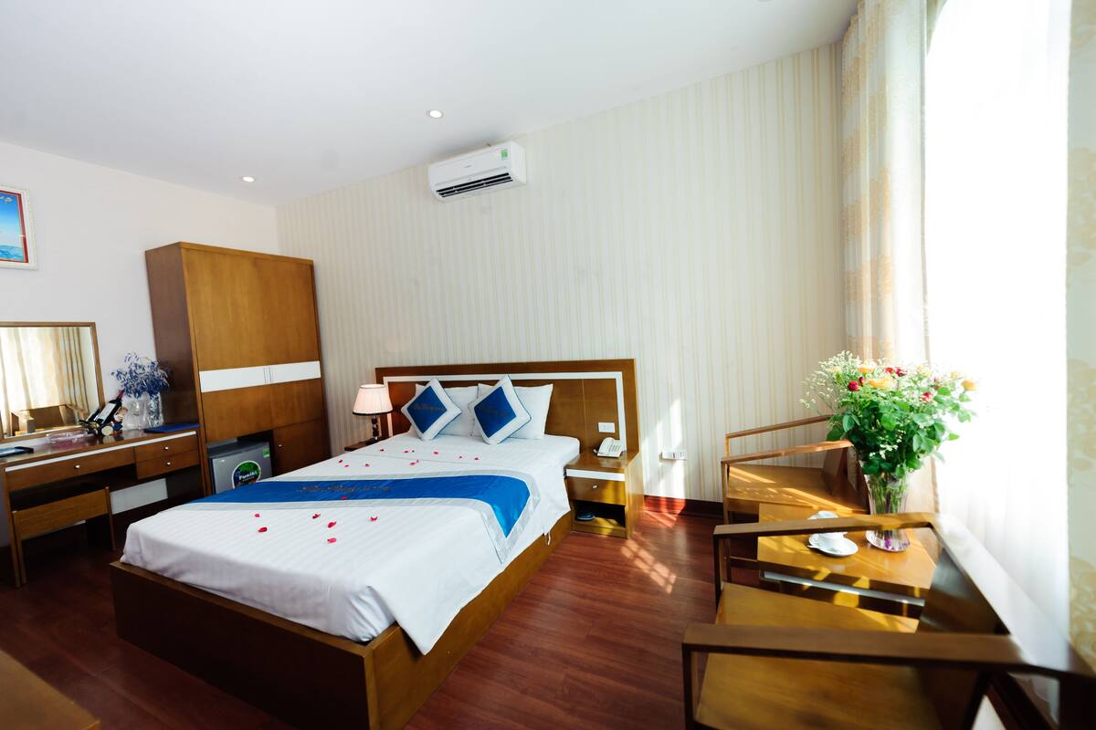 A hotel room with a wooden bed, a desk, a chair, a nightstand, and a vase with flowers.