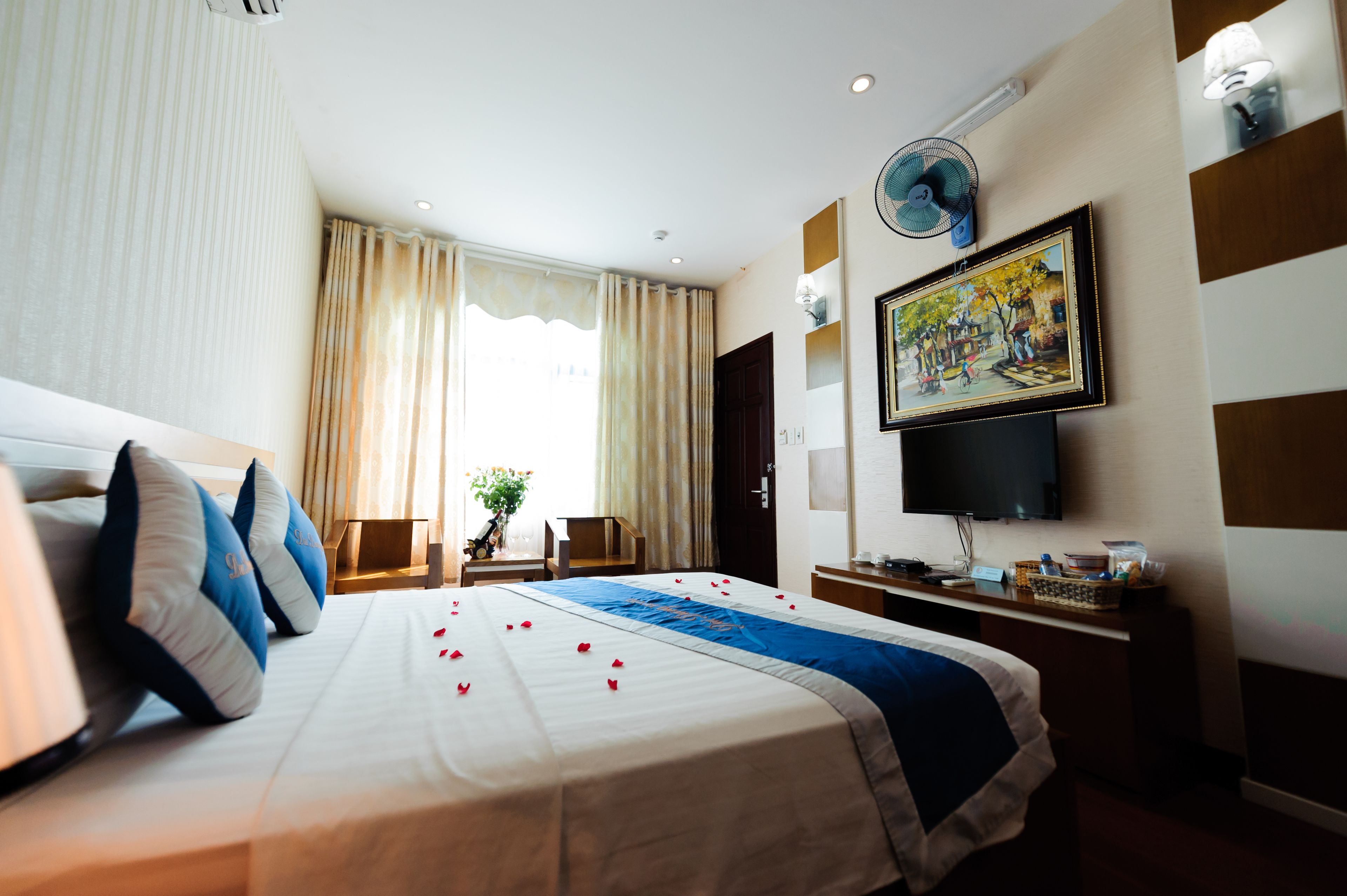 A hotel room with a large bed, a flat-screen TV, a desk with a lamp, a fan, and a painting on the wall.