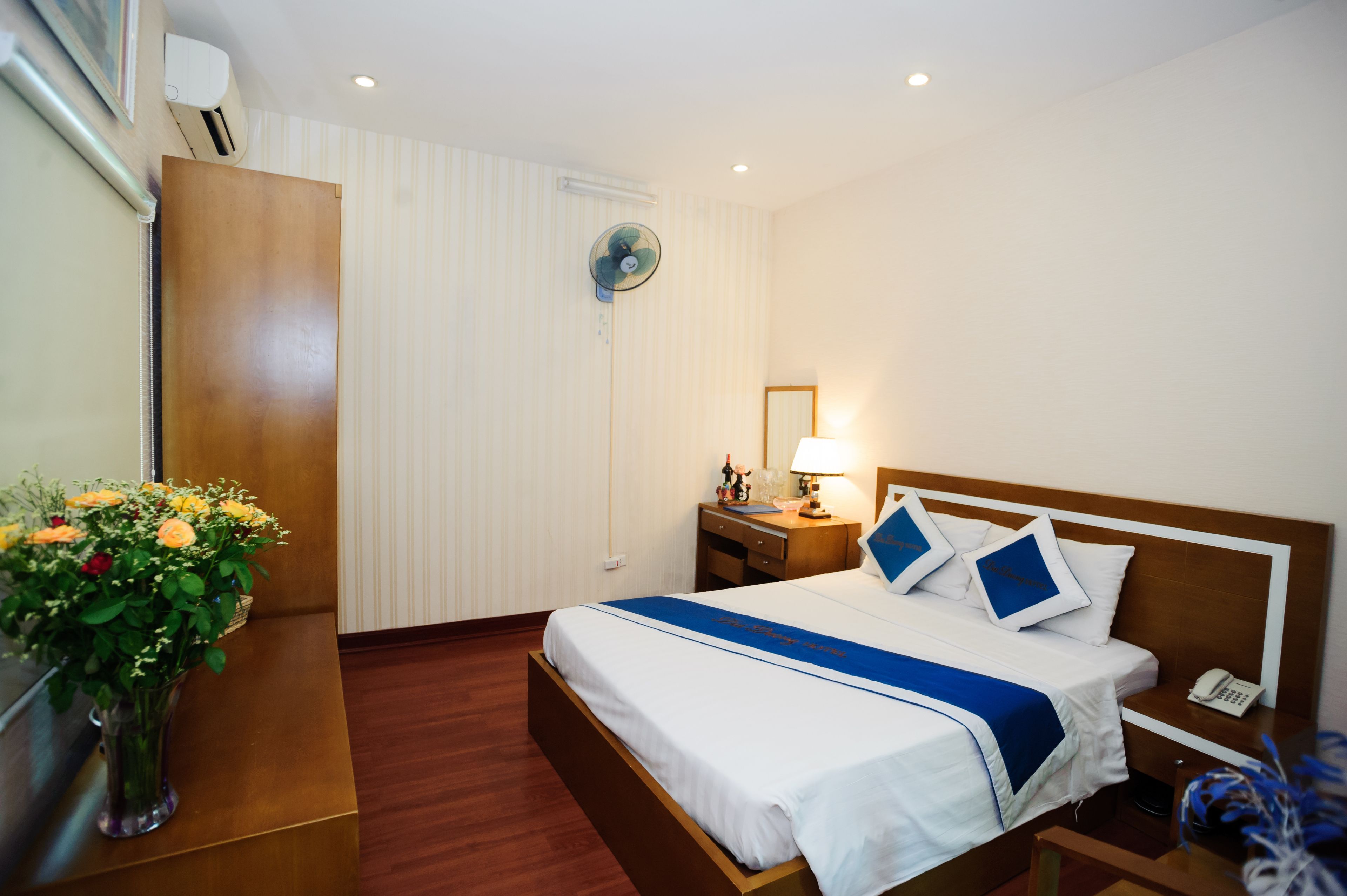 A hotel room with a bed, bedside table, lamp, and a vase with flowers.