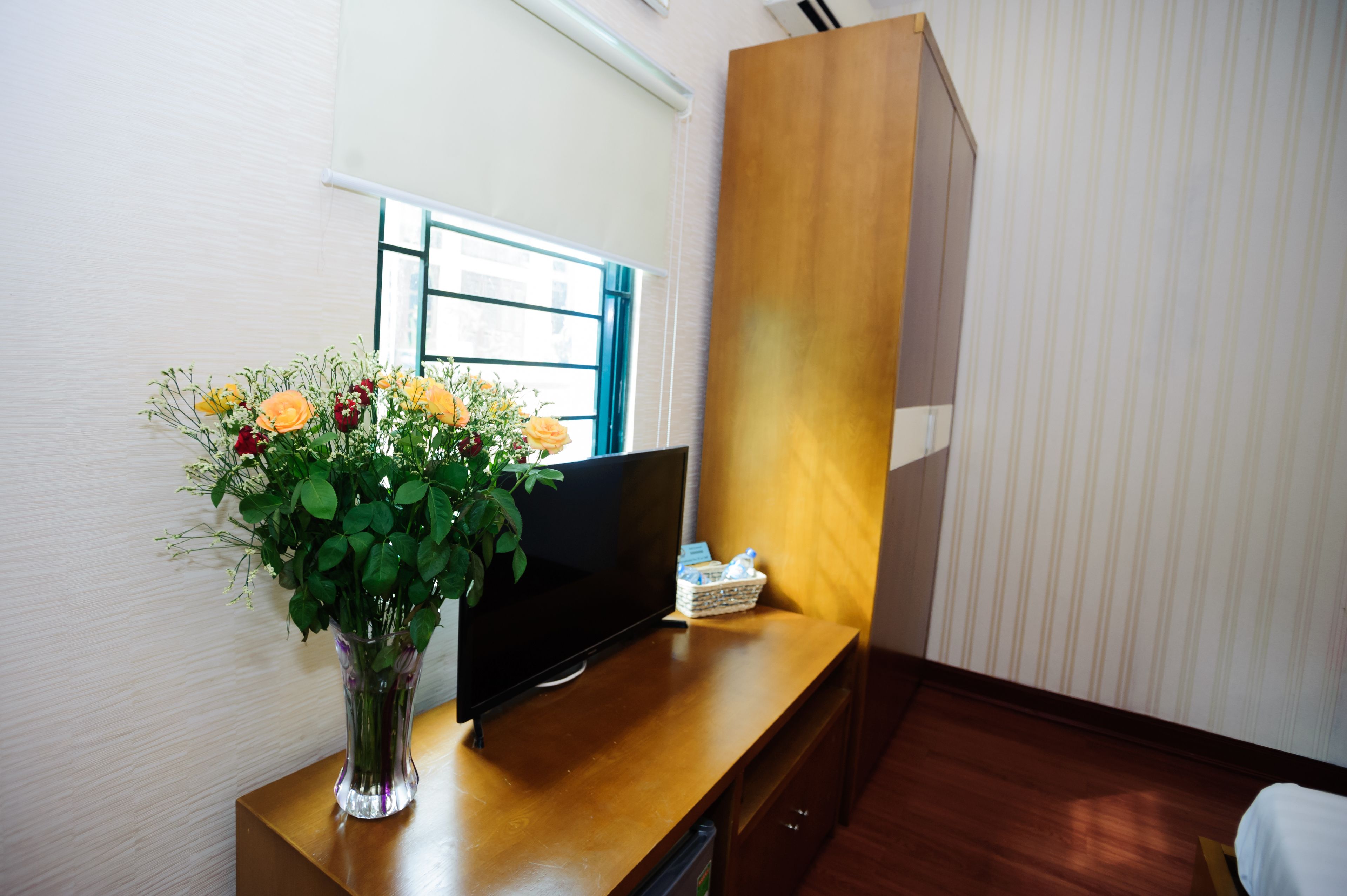 Et værelse med et træskrivebord, et fladskærms-tv, en vase med blomster og et vindue med persienner.