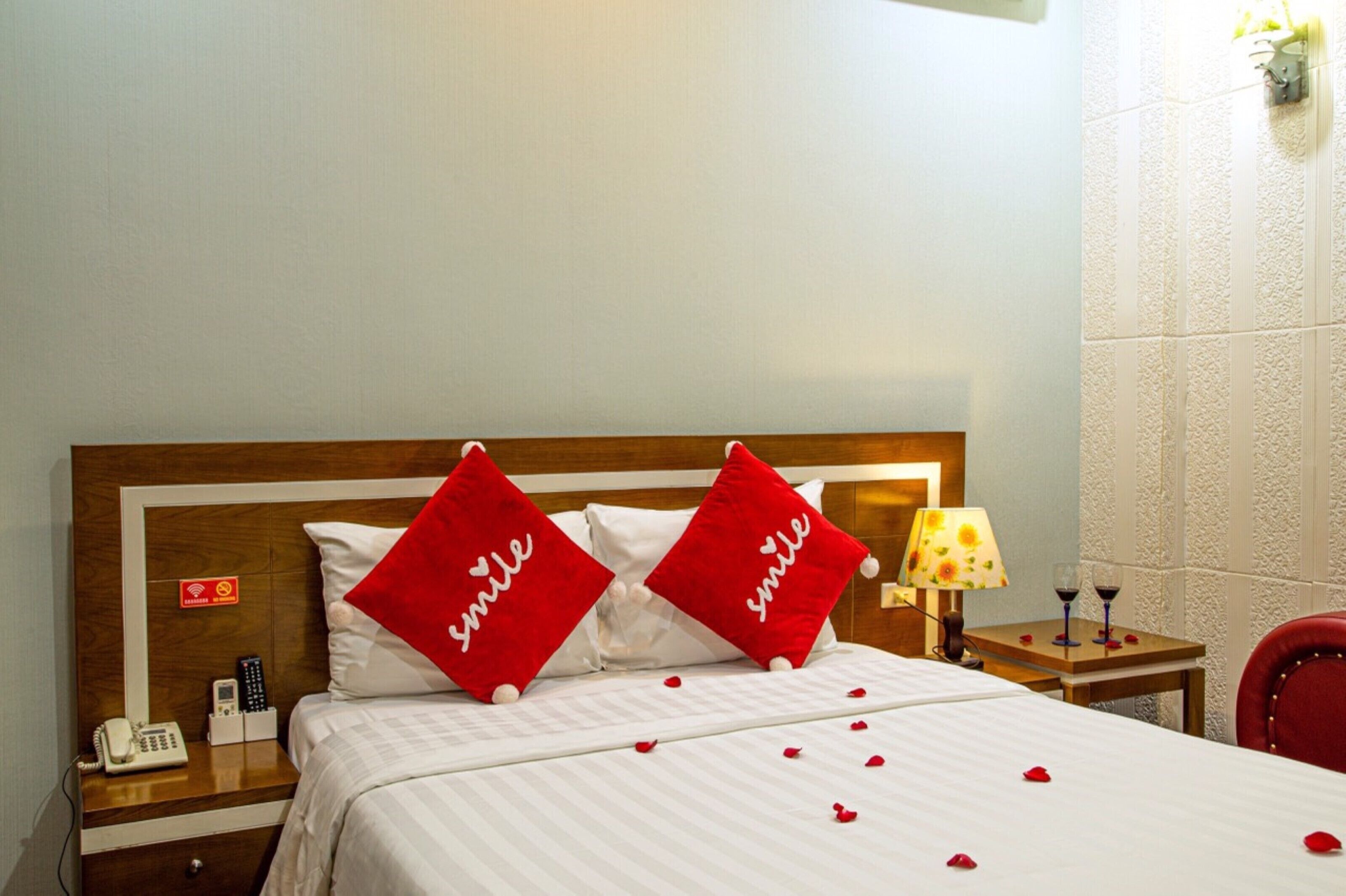 Habitación de hotel con una cama, dos almohadas rojas con la palabra “smile” y una mesita de noche con una lámpara, un teléfono, y una pequeña mesa con copas de vino y pétalos de rosa.