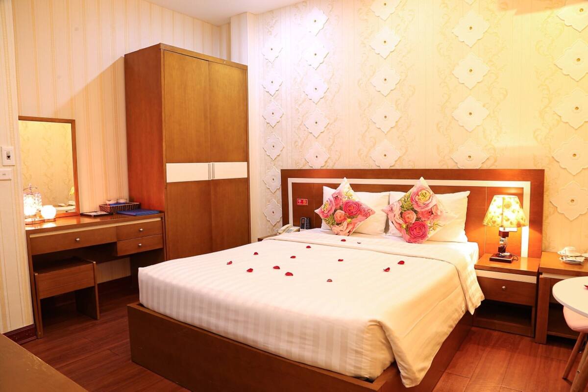 A hotel room with a large bed, wooden wardrobe, bedside table with lamp, and a desk with a mirror.
