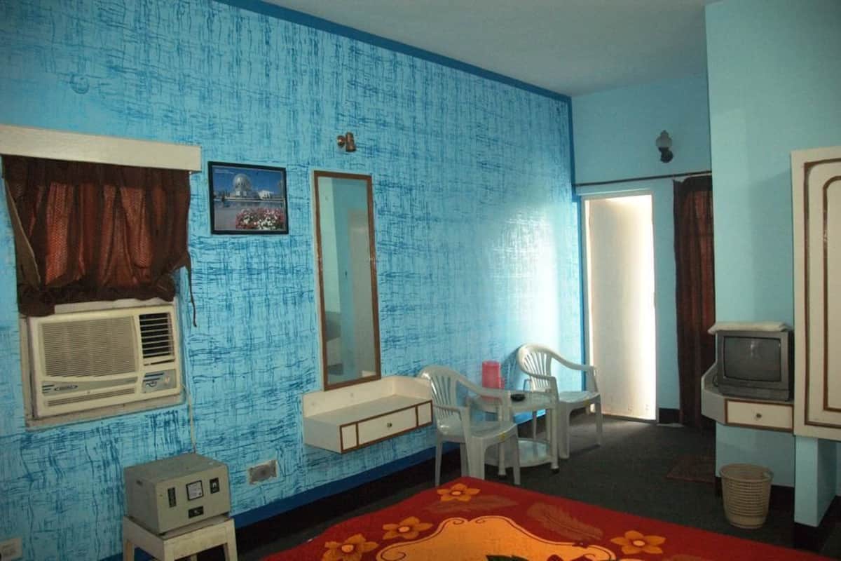 Una habitación con pared azul con textura, una cama con colcha floral roja, una pequeña mesa con sillas, un televisor y un equipo de aire acondicionado.