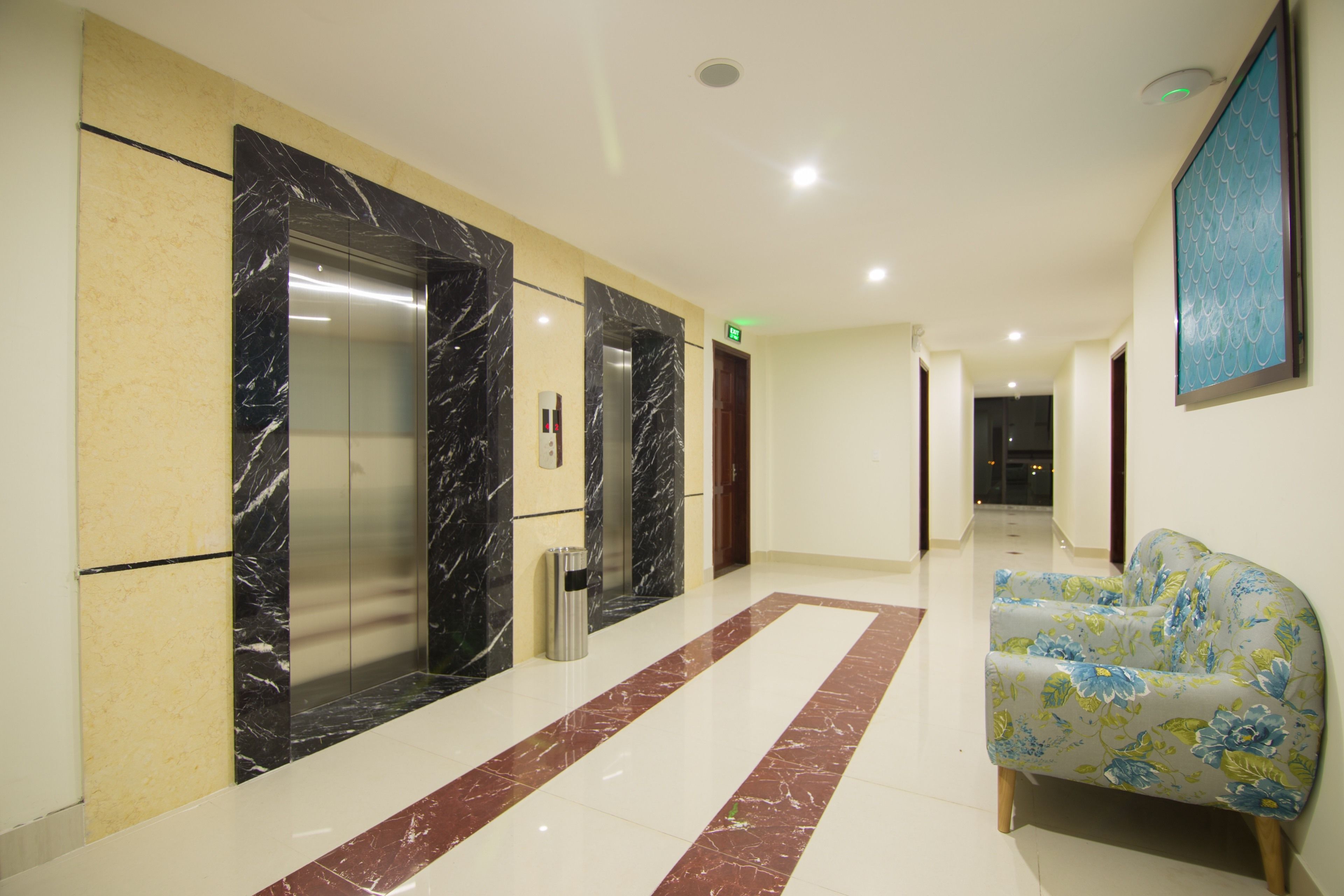 A hotel lobby with marble flooring, two elevators, a trash bin, and a floral sofa.