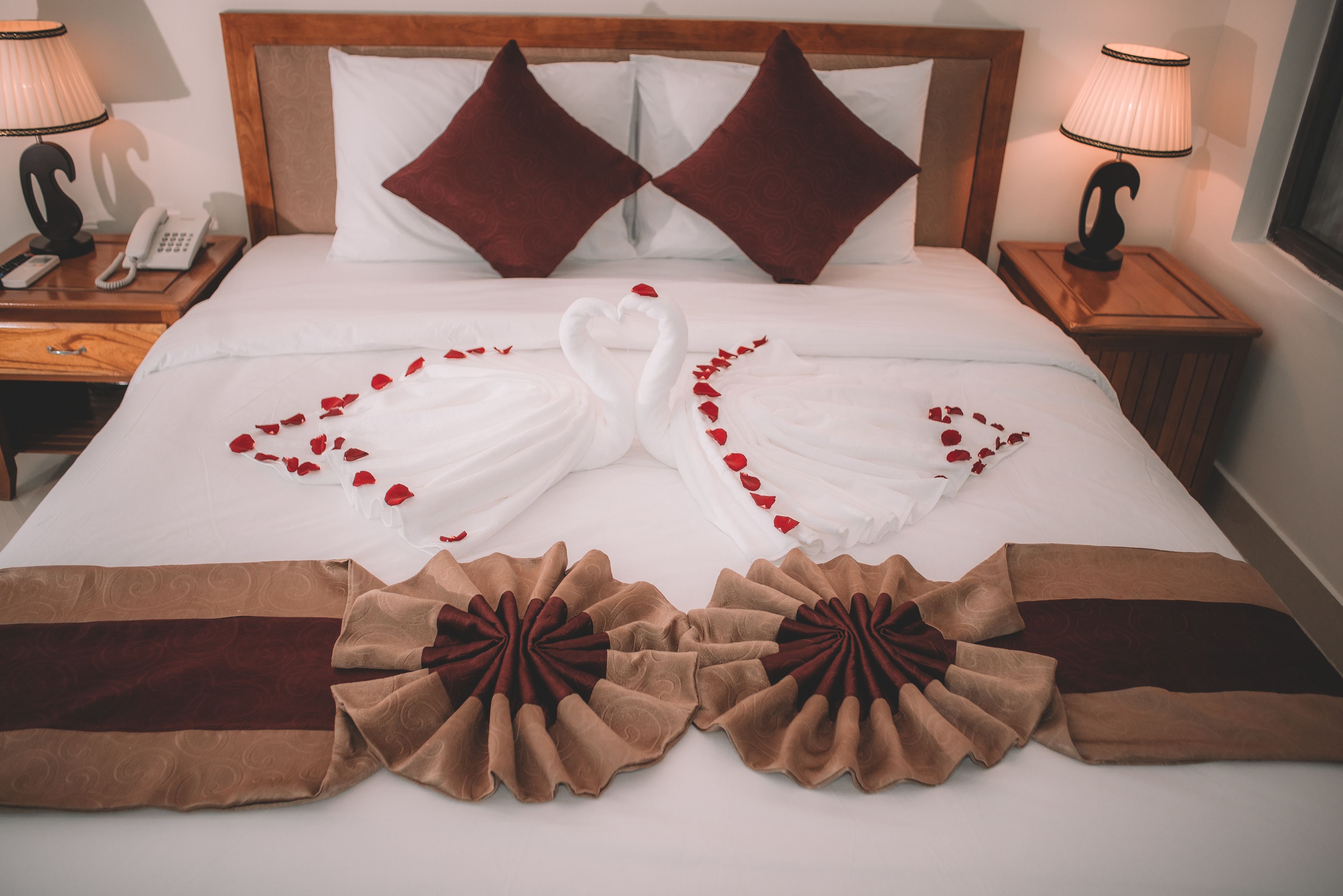 Un lit bien fait, agrémenté d’une décoration en forme de cœur réalisée avec des serviettes et des pétales de rose, de deux oreillers marron foncé et d’une tête de lit en bois.