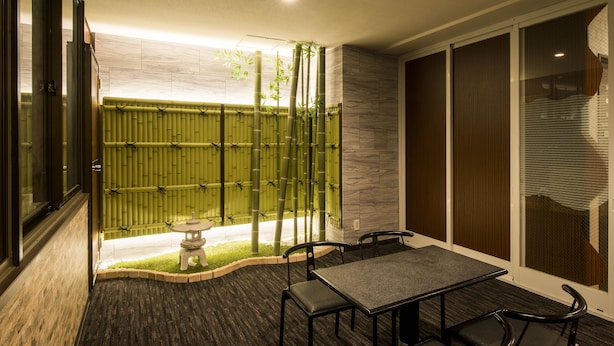 A modern interior with a dining area featuring a table and chairs, a small garden with a bamboo fence, and a view through large windows. - Sejour Inn Capsule (Hiroshima)