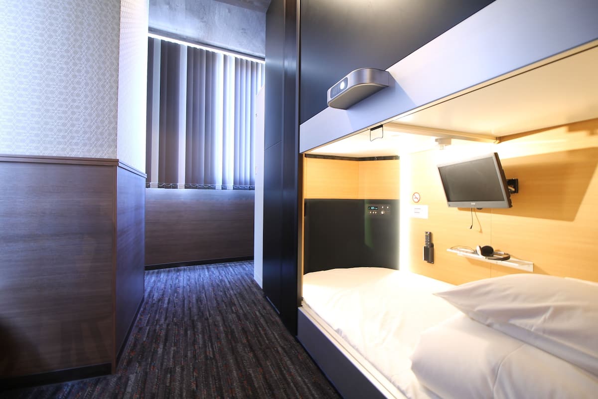 A hotel room with a bed, a TV mounted on the wall, a window with blinds, and a carpeted floor.