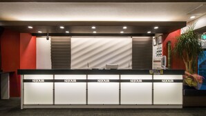 A reception desk with 'SEJOUR' signs, a plant, and a wall with a textured pattern. - Sejour Inn Capsule (Hiroshima)
