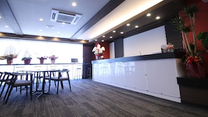 A reception area with a counter, chairs, potted plants, and a view through the window. - Sejour Inn Capsule (Hiroshima)